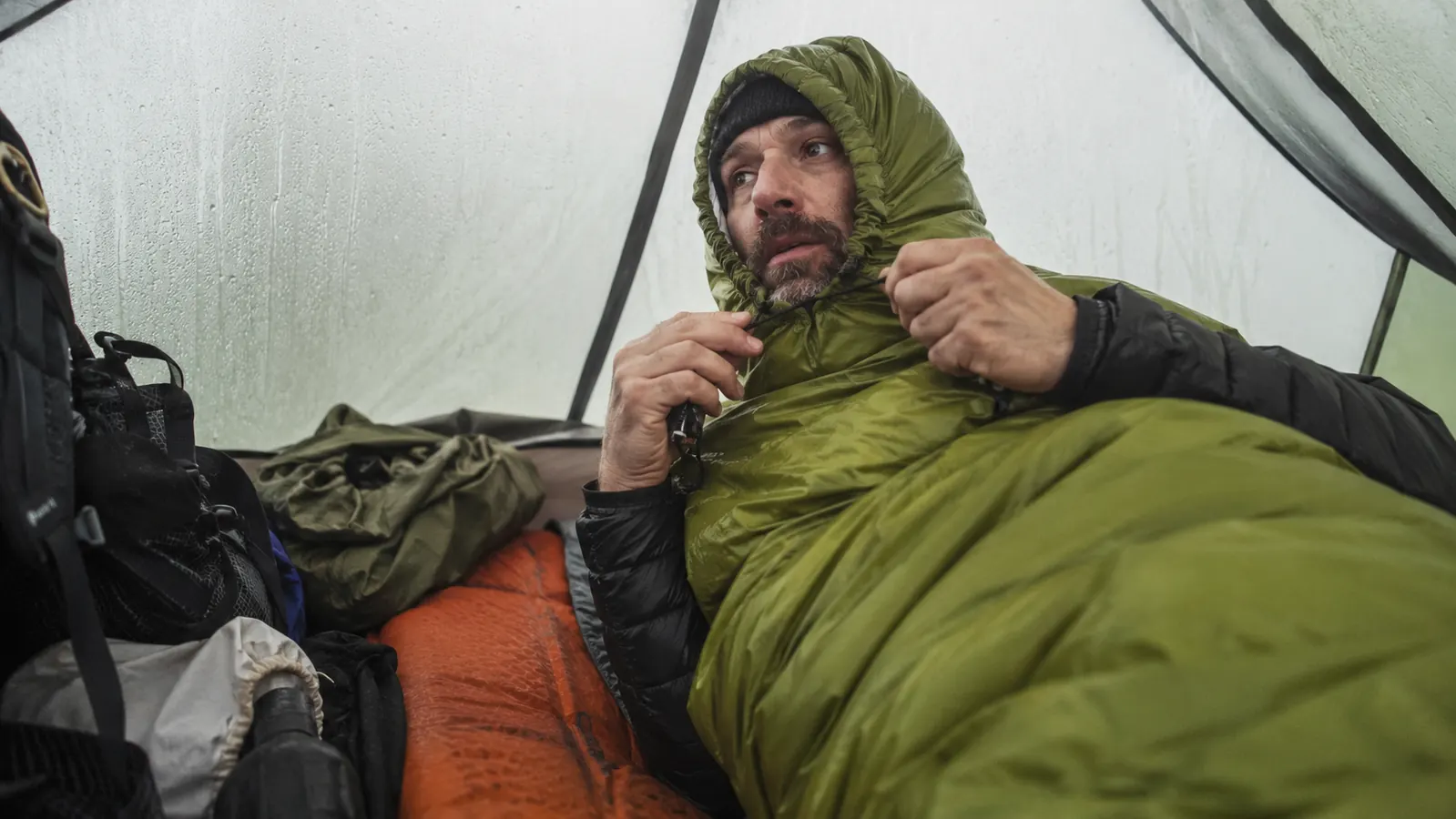 Camper zipping up a mummy sleeping bag inside a tent with frost visible on the tent walls