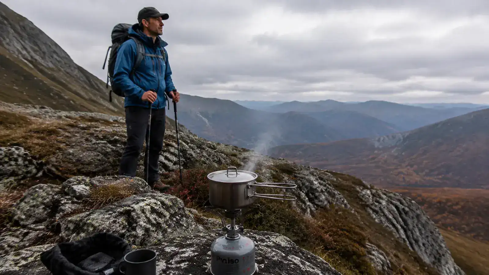 Titanium backpacking stove with pot on a rocky ridgeline, hiker in background looking at a map