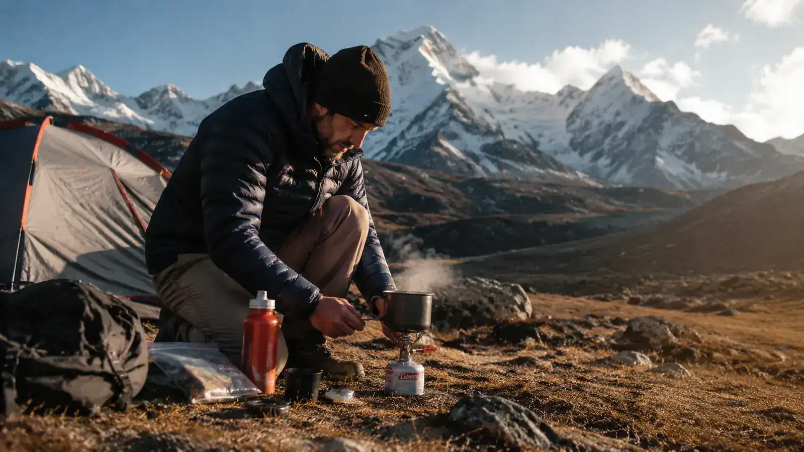 Hiker boiling water on a canister stove at a mountain campsite with snow-capped peaks in the distance