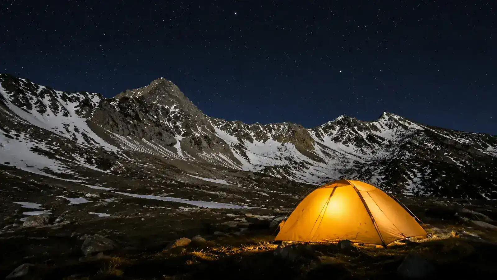 Campsite in the mountains at night, tent glowing amber from inside, stars visible, snow on ridgeline in background