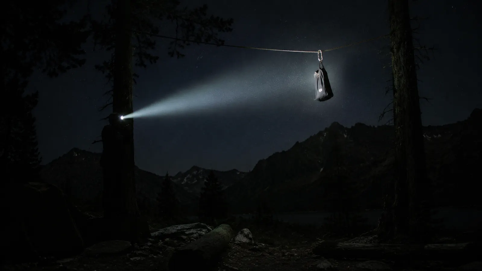Night shot of a campsite with a headlamp beam cutting through the dark toward a food bag hanging from a bear line