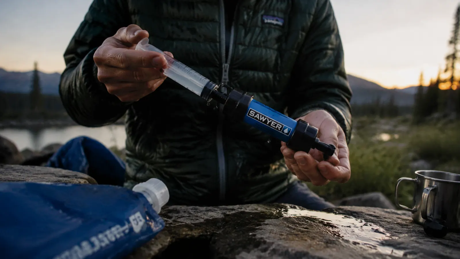 Backpacker using a syringe to backflush a Sawyer Mini filter at a campsite, clean water flowing through onto dry leaves