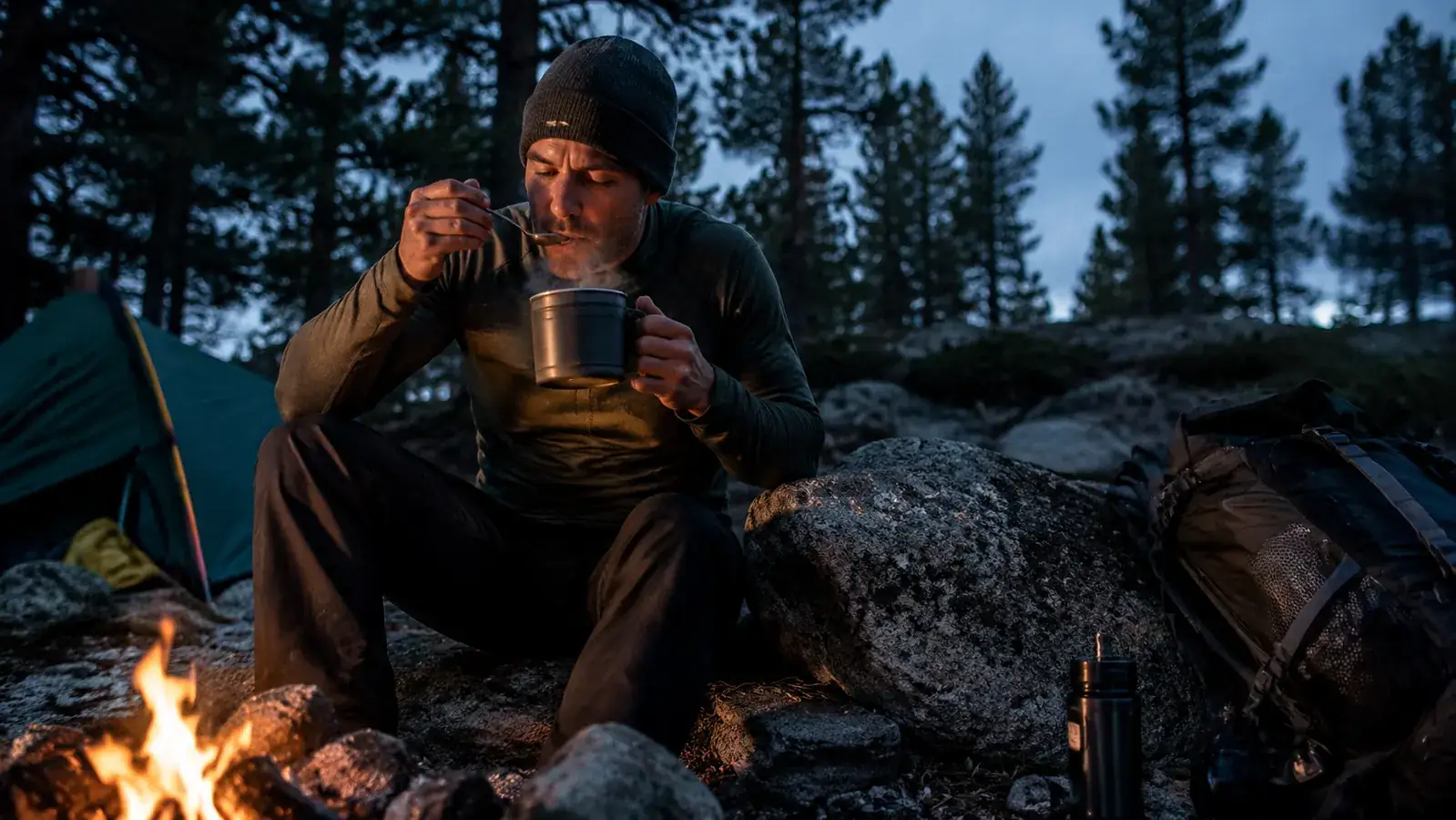 Person wearing a base layer and wool hat eating a hot camp meal before bed at a campsite fire