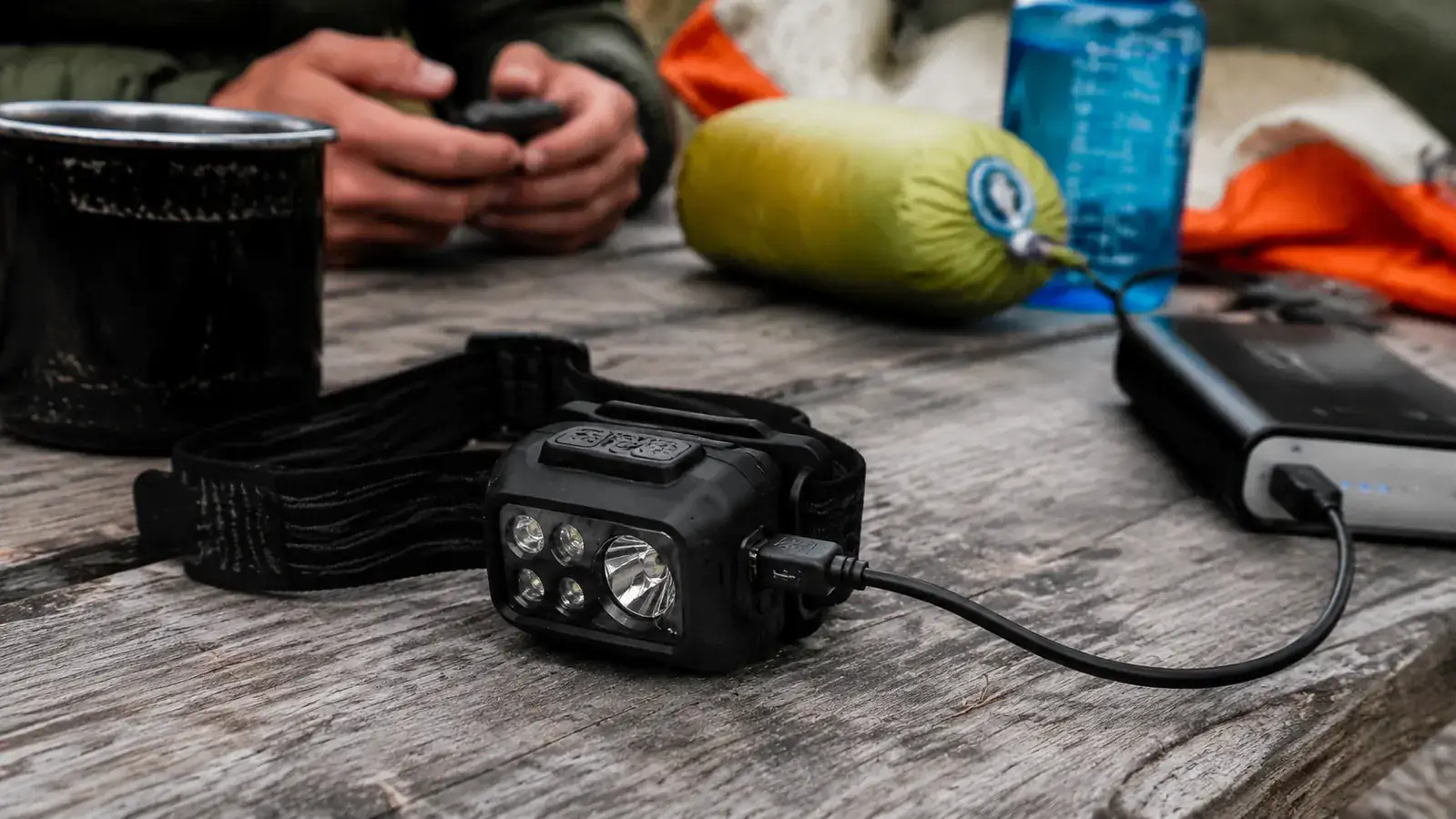 LHKNL headlamp showing USB charging port with a charging cable plugged in, resting on a picnic table next to a camp lantern