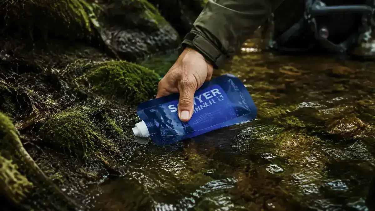 Hand holding Sawyer Mini filter while filling a pouch from a shallow stream