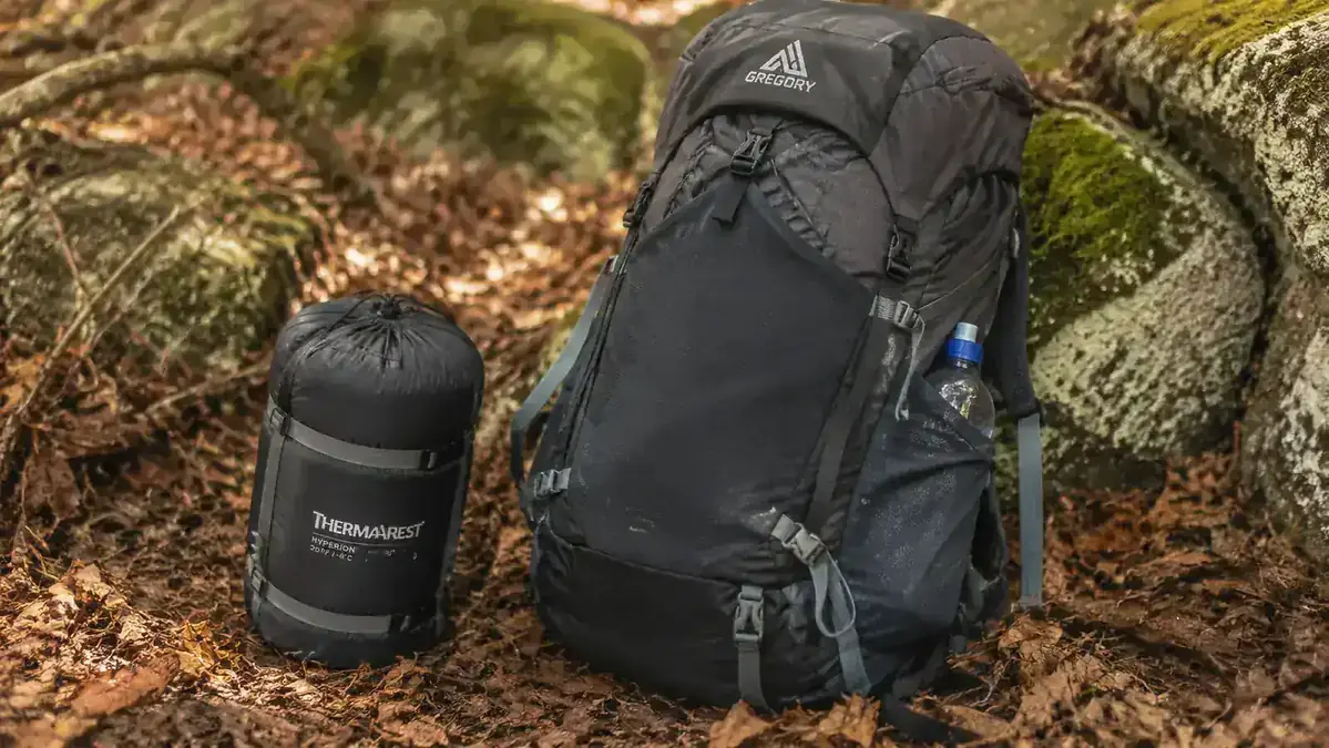 Teton Sports Celsius mummy bag stuffed into its compression sack, sitting next to a daypack on a forest floor