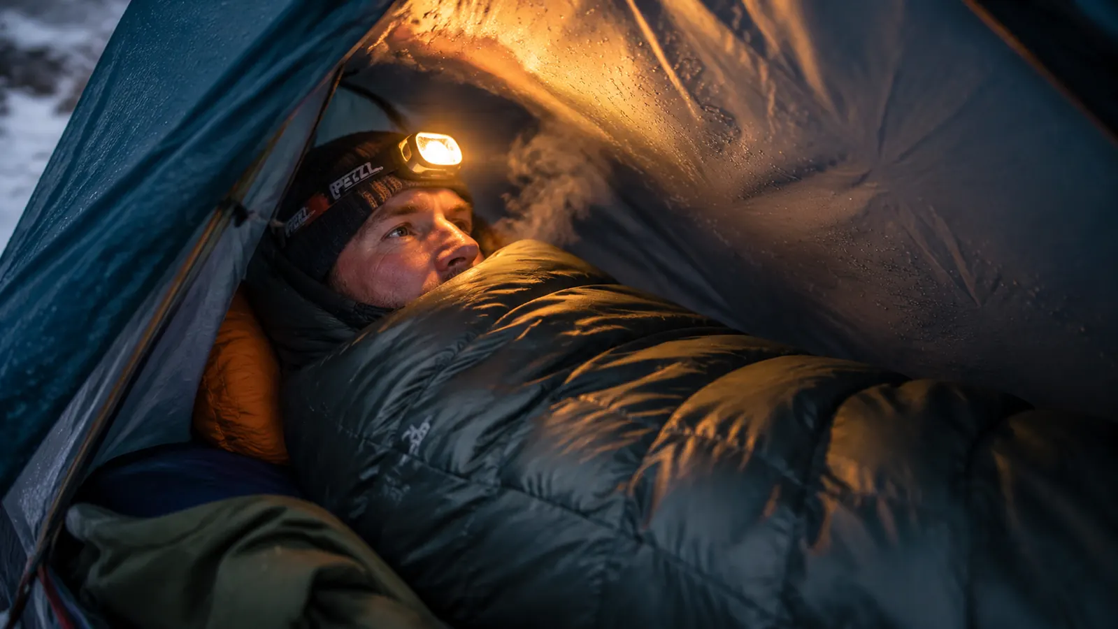 Camper inside a mummy sleeping bag inside a tent, head visible above the bag cinched snug, condensation on tent wall