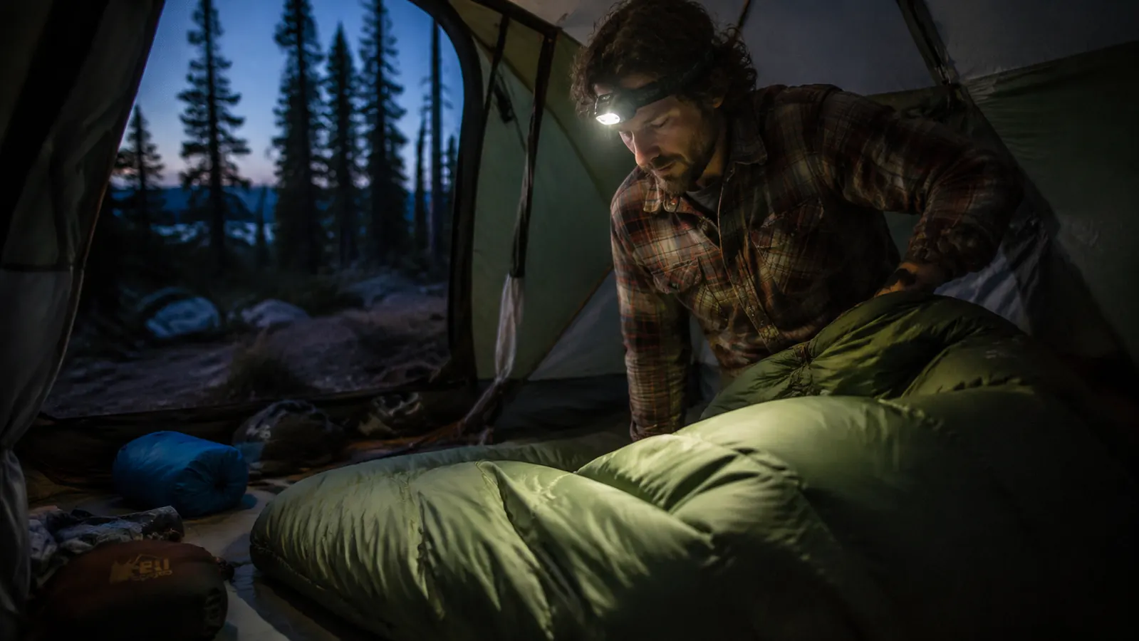 Person sliding into the Teton Sports Celsius mummy bag at a car-camping site, headlamp on, trees in background at dusk
